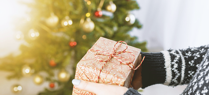 Hand hält ein Weihnachtsgeschenk und im Hintergrund ist ein geschmückter Weihnachtsbaum